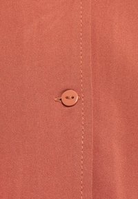 Koraalrood stof met een gladde textuur heeft een knoopdetail en contrasterend stiksel langs de naad.