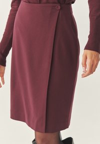 Burgundská sukňa na ceruzku vyrobená z hladkej látky, s bočným gombíkom a jemným záhybom, s dĺžkou po kolená a minimalistickým dizajnom.