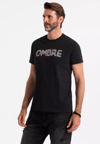 Sort bomulds t-shirt med "OMBRE" tekst i et stribet design. Korte ærmer og rund halsudskæring. Bæres med slidte sorte jeans.