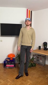 Μπεζ πλεκτό πουλόβερ με V-λαιμόκοψη και μακριά μανίκια, συνδυασμένο με σκουρόχρωμα γκρι τζιν με καμπάνες και γυαλιστερά καφέ παπούτσια. Το φόντο περιλαμβάνει τηλεόραση και διακοσμητικά.