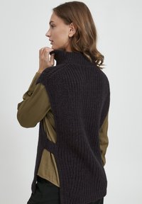 Mørk lilla strikket ærmeløs sweater med høj krave og sideskæringer, lagdelt over en grøn bluse med samlet ærmer.