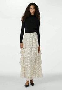 Camisola preta de mangas compridas e uma saia maxi creme fluida com três camadas de babados e bolinhas pretas. Sapatos pretos planos completam o visual.