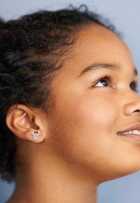 Orecchini con borchie che presentano un design di Topolino con un fiocco rosso, in tonalità argento e accenti scintillanti, indossati da una persona con capelli ricci.