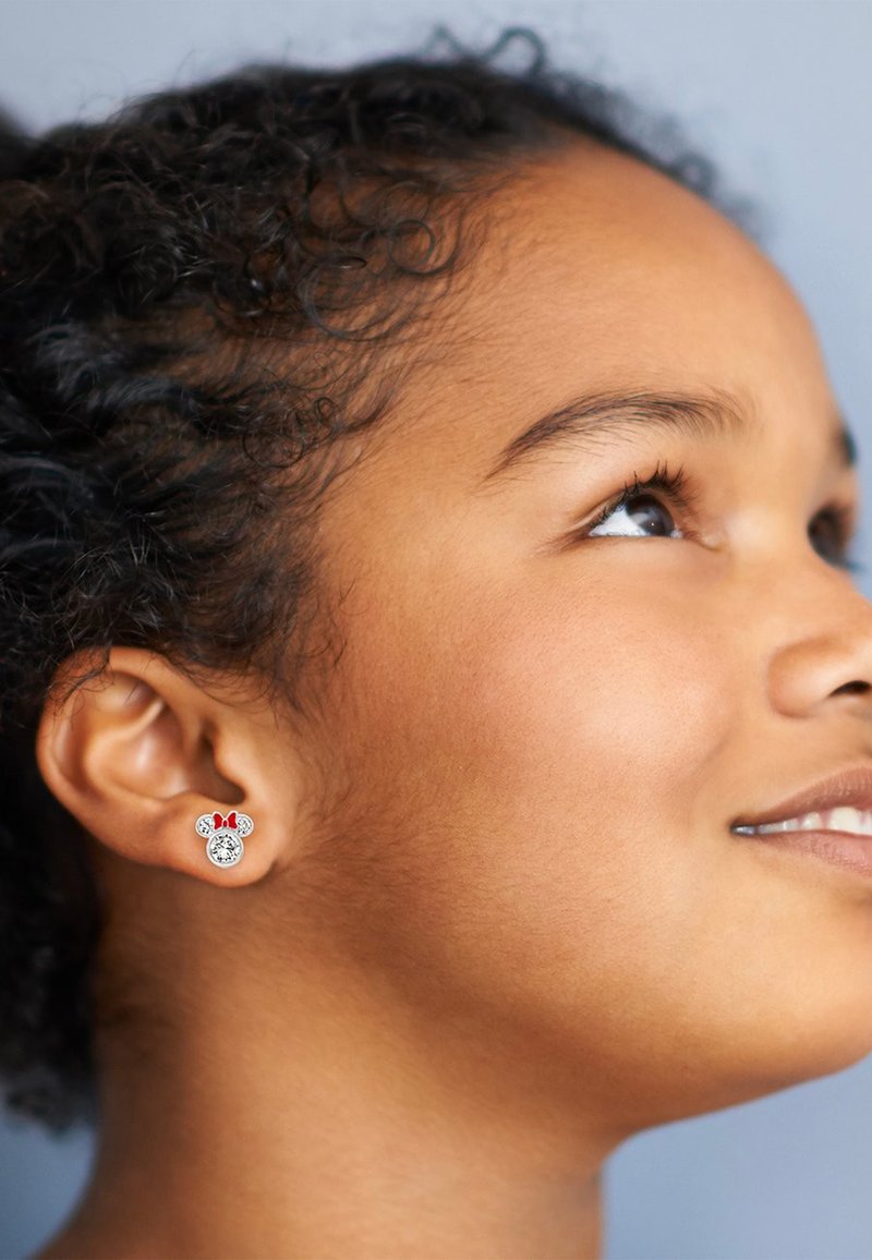 Orecchini con borchie che presentano un design di Topolino con un fiocco rosso, in tonalità argento e accenti scintillanti, indossati da una persona con capelli ricci.