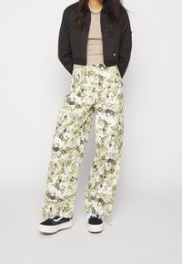 Svart kort jacka, beige topp, löst sittande byxor med grönt blommönster, vita och bruna sneakers med svarta detaljer.