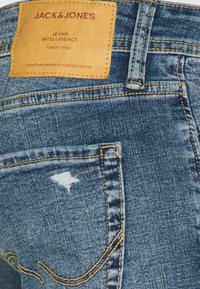 Denimske hlače v srednji modri barvi, s kožnim etiketo blagovne znamke, poudarki v šivu in obrabljenim detajlom na zadnjem žepu.