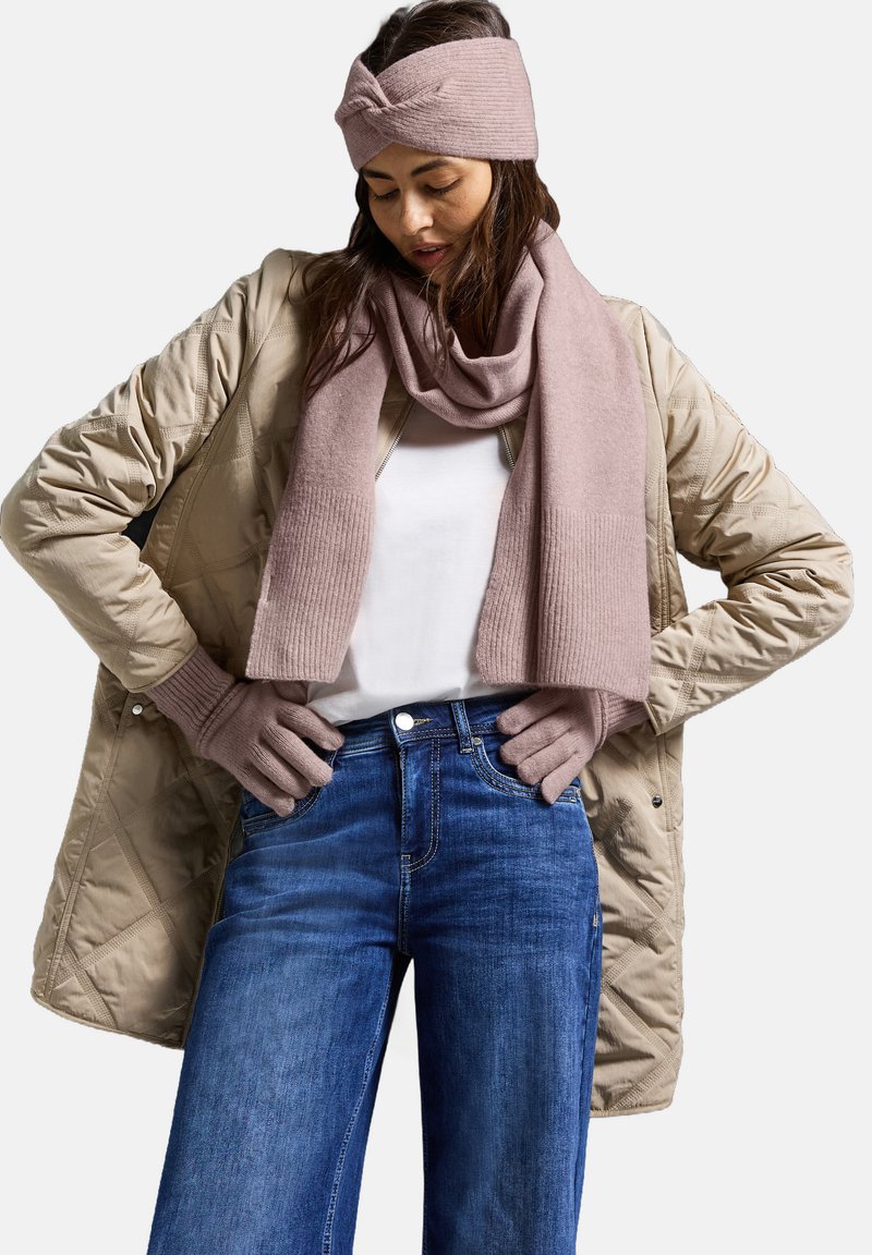 Beige kviltad kappa, ljusrosa ribbad scarf och huvudband, mörkblå jeans och rosa handskar, som visar en avslappnad lager-på-lager-outfit.