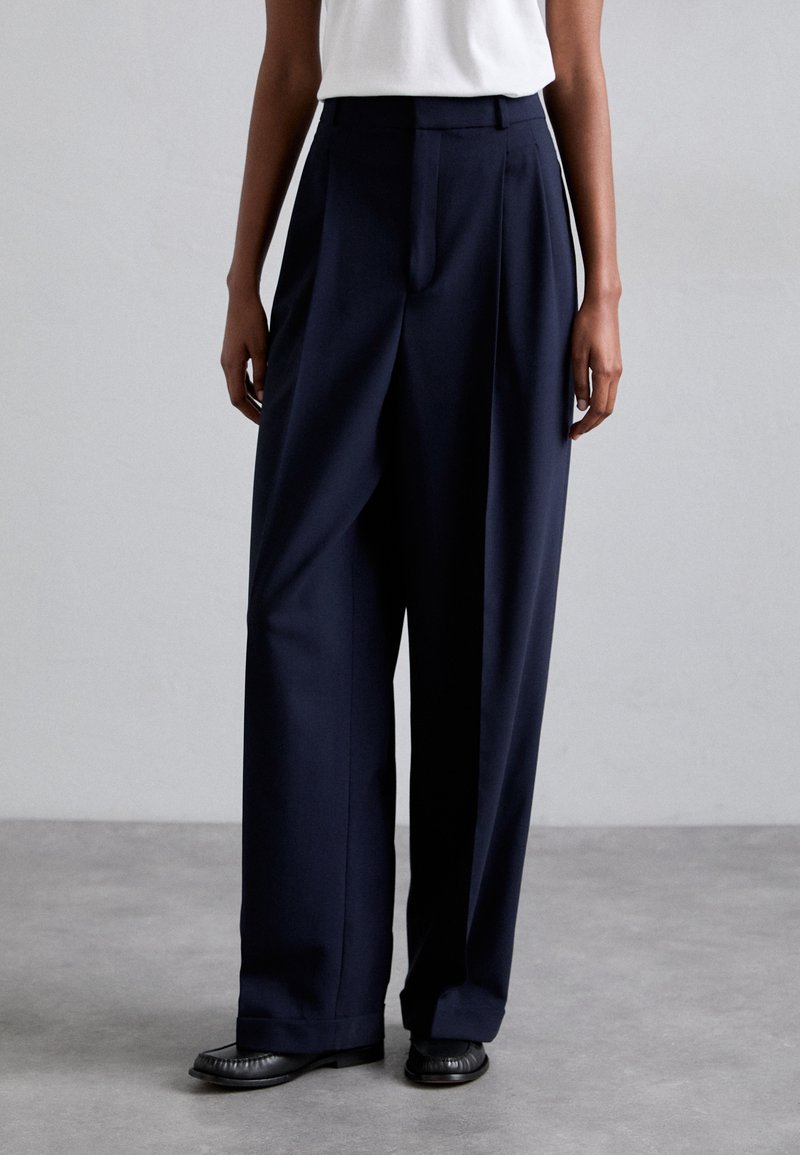 Navy wide-leg trousers made of a smooth fabric, featuring pleats and a tailored waist, paired with flat black shoes.