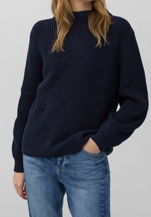 Personne portant un pull en tricot bleu foncé et un jean bleu clair, avec une main dans la poche sur un fond uni.