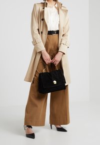 Czarna zamszowa torebka na złotym zapięciu, trzymana w jednej ręce. Noszona z beżowym płaszczem trenczowym, białą koszulą i brązowymi spodniami o szerokich nogawkach.