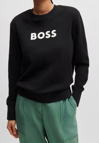 Černá mikina s bílým logem "BOSS". Má kulatý výstřih a dlouhé rukávy, v kombinaci se světle zelenými cargo kalhotami. Měkká struktura látky.