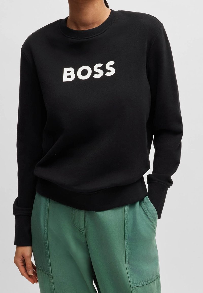 Černá mikina s bílým logem "BOSS". Má kulatý výstřih a dlouhé rukávy, v kombinaci se světle zelenými cargo kalhotami. Měkká struktura látky.