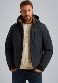 Chaqueta acolchada negra con capucha, que cuenta con una cremallera frontal, dos bolsillos en el pecho y un forro amarillo en contraste, llevada sobre un sudadera beige.