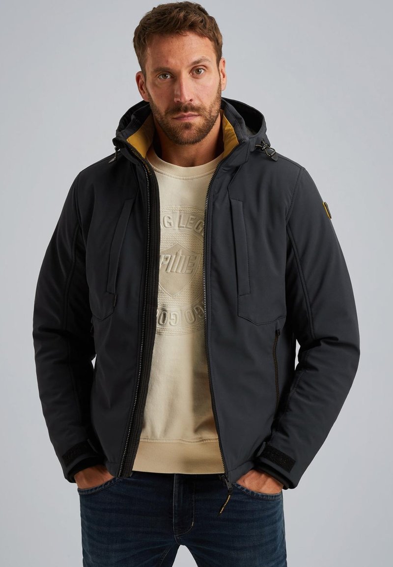 Chaqueta acolchada negra con capucha, que cuenta con una cremallera frontal, dos bolsillos en el pecho y un forro amarillo en contraste, llevada sobre un sudadera beige.
