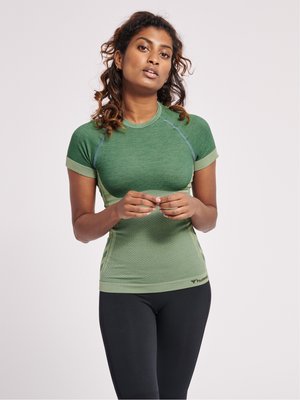 Vrouw met krullend haar die een groen sporttopje met korte mouwen en zwarte leggings draagt, staat met de handen zachtjes gesloten.