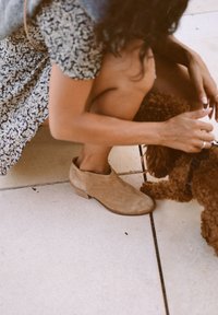 Botas de ante hasta el tobillo en color marrón claro, con puntera redondeada y tacón bajo, que lleva una persona de rodillas al lado de un perro pequeño sobre una superficie de baldosas.