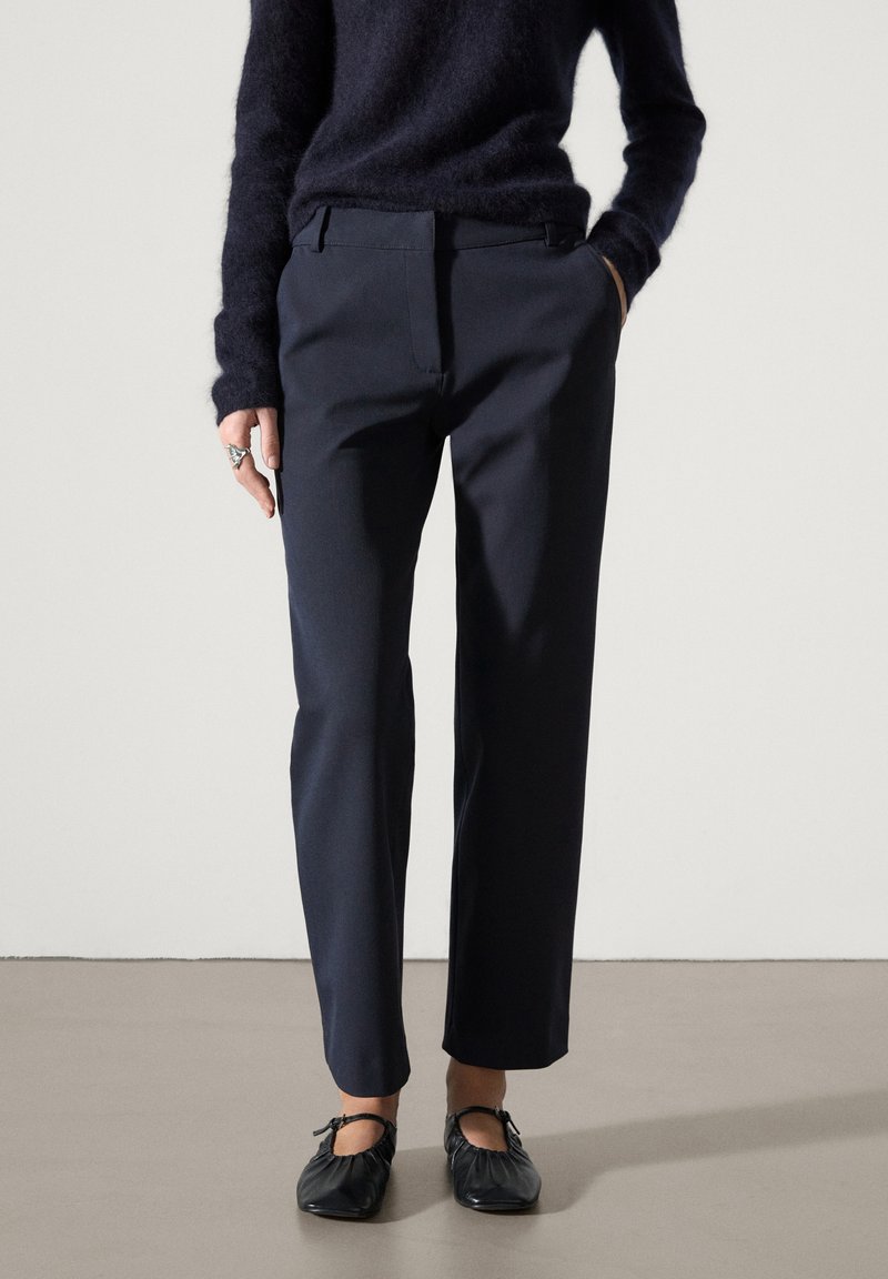 Femme portant un pantalon droit bleu marine foncé, un pull foncé assorti et des ballerines noires, debout sur un sol neutre.