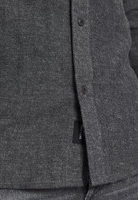 Camisa de patrón de espiga gris con botones negros, cuello puntiagudo y etiqueta lateral. Tejido texturizado con un sutil brillo.