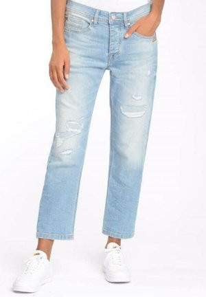 Osoba ubrana w jasnoniebieskie podarte dżinsy i białe trampki, stojąca z jedną ręką w kieszeni na białym tle.