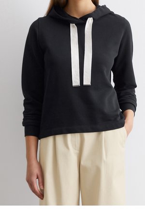 Sweat à capuche noir avec larges cordons blancs porté avec un pantalon beige taille haute, mains détendues, cadrage raccourci du cou aux hanches.