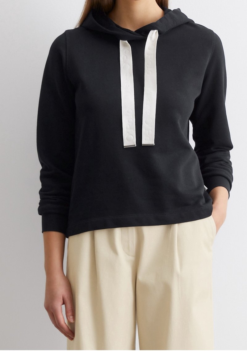 Sweat à capuche noir avec larges cordons blancs porté avec un pantalon beige taille haute, mains détendues, cadrage raccourci du cou aux hanches.