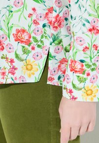 Närbild av en hand vid fållen och ärmslutet på en vit blommig skjorta med röda, rosa och gula blommor, tillsammans med gröna byxor.