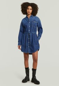 Τζιν φόρεμα με μακριά μανίκια, κουμπιά μπροστά και ζώνη στη μέση; διαθέτει τσέπη στο στήθος και καμπύλη ραφή στο στρίφωμα. Φορεμένο με μαύρες μπότες μέχρι τον αστράγαλο.