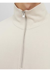 Sudadera cremosa de cremallera con cuello alto, cremallera de metal y tejido suave y texturizado. Los detalles de costura añaden interés visual.