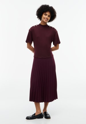 Femme aux cheveux bouclés portant un haut bordeaux à manches courtes avec boutons aux épaules et une jupe midi plissée assortie avec des mocassins noirs.