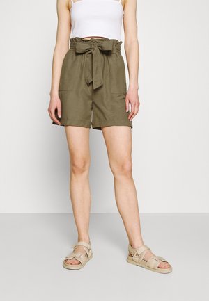 Kvinde iført olivengrønne shorts med høj talje og bindebælte, hvid crop top og beige åbne sandaler stående på gråt gulv.