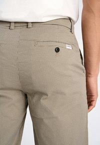 Pantalones beige con estampado que presentan una superficie texturizada, un bolsillo trasero con un botón negro y una pequeña etiqueta en la cintura.