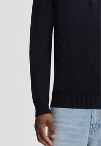 Marineblauer Strickpullover mit gerippten Bündchen und Saum, ausgestattet mit einem runden Ausschnitt. Die Hand liegt auf dem Bund einer hellblauen Jeans.