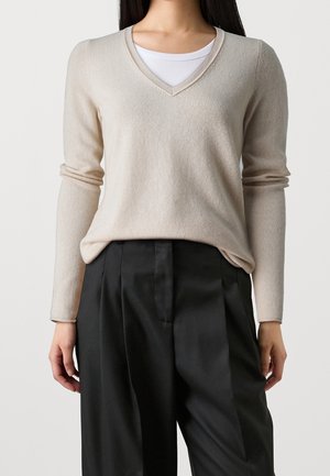 Femme portant un pull beige à col en V sur un débardeur blanc, associé à un pantalon noir plissé taille haute.