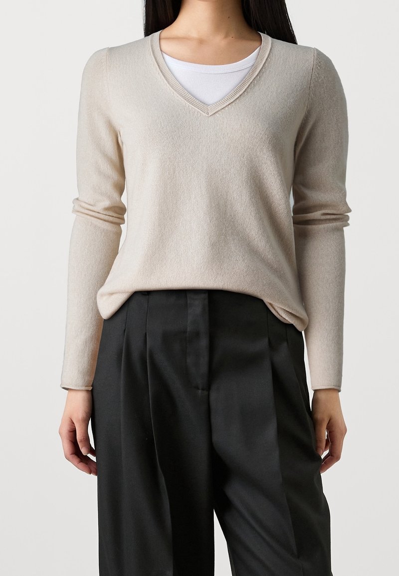 Femme portant un pull beige à col en V sur un débardeur blanc, associé à un pantalon noir plissé taille haute.