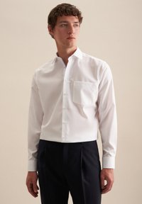 Camicia bianca a maniche lunghe con bottoni, realizzata in tessuto morbido, dotata di taschino sul petto, colletto classico e vestibilità su misura, abbinata a pantaloni scuri.