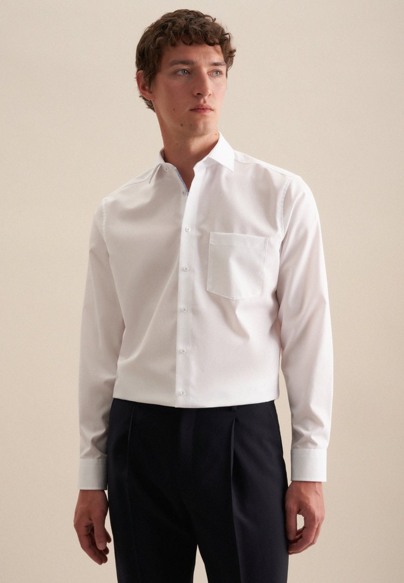 Camicia bianca a maniche lunghe con bottoni, realizzata in tessuto morbido, dotata di taschino sul petto, colletto classico e vestibilità su misura, abbinata a pantaloni scuri.