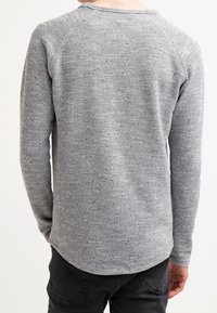 Osoba ubrana w szary, długi sweter dzianinowy i czarne spodnie, pokazana od tyłu na jednolitym jasnym tle.