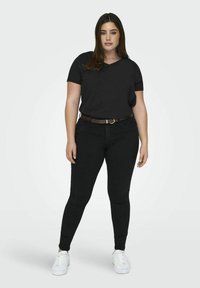 T-shirt noir à manches courtes associé à un jean skinny noir. Le modèle porte une ceinture marron, des baskets blanches et arbore des cheveux raides et sombres.