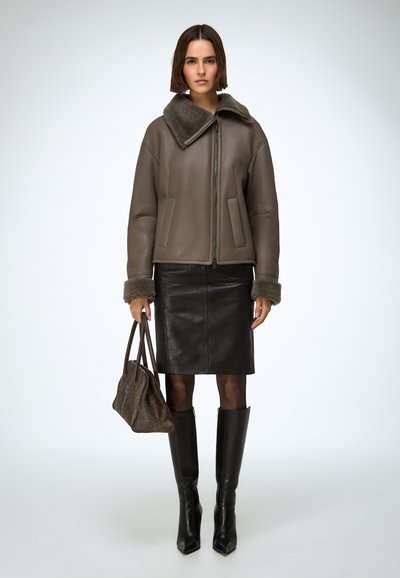 Hnedá kožená bomber bunda s kožušinovým golierom, v kombinácii s čiernou ceruzkovou sukňou, čiernymi čižmami na vysokom opätku po kolená a sivou kabelkou.