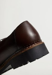 Talons de chaussures en cuir brun foncé texturé avec semelle en caoutchouc noir épaisse et coutures détaillées sur fond clair uni.