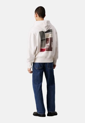 Sudadera blanca con un diseño gráfico en la parte trasera que presenta formas abstractas en gris y rojo. Combinada con jeans de denim azul sueltos y zapatos negros.