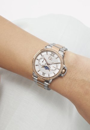 Polshorloge met zilveren en roségouden metalen band, witte wijzerplaat, Romeinse cijfers, subwijzers voor dag en datum, en maanfase-indicator, gedragen om de pols.