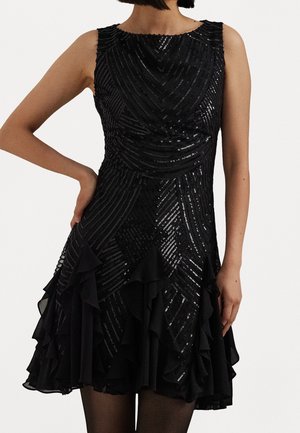 Sleeveless black dress with diagonal sequin stripes and layered ruffles on the skirt, worn by a person with one hand on hip.