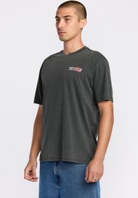 T-shirt en coton gris foncé à manches courtes, avec un petit logo sur la poitrine en rouge et blanc. Col rond, coupe décontractée.