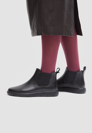 Stivaletti in pelle nera indossati con calze bordeaux e gonna scura su uno sfondo chiaro.