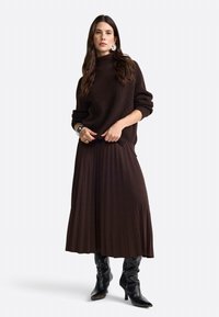 Brauner gerippter Pullover mit hohem Kragen und oversize Schnitt, kombiniert mit einem plissierten dunkelbraunen Rock und schwarzen kniehohen Stiefeln mit Absatz.