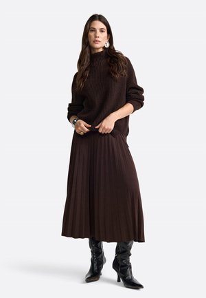 Maglione marrone a coste con colletto alto e vestibilità oversize, abbinato a una gonna plissettata marrone scuro e stivaletti neri con tacco al ginocchio.