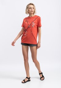 Rode katoen t-shirt met abstracte grafische afbeeldingen en een losse pasvorm, gecombineerd met donkere shorts en zwarte sandalen met bandjes op een effen achtergrond.