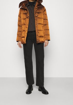 Vrouw draagt een glanzend koperen donsjack met kap, een zwart button-up overhemd, zwarte broek en zwarte enkellaarsjes, staand op een grijze vloer.