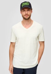 Helles Cremefarbenes V-Ausschnitt-T-Shirt mit kurzen Ärmeln, aus einem strukturierten Material. Kombiniert mit einer marineblauen Cap mit einem grünen "PURE JUICE" Grafik.